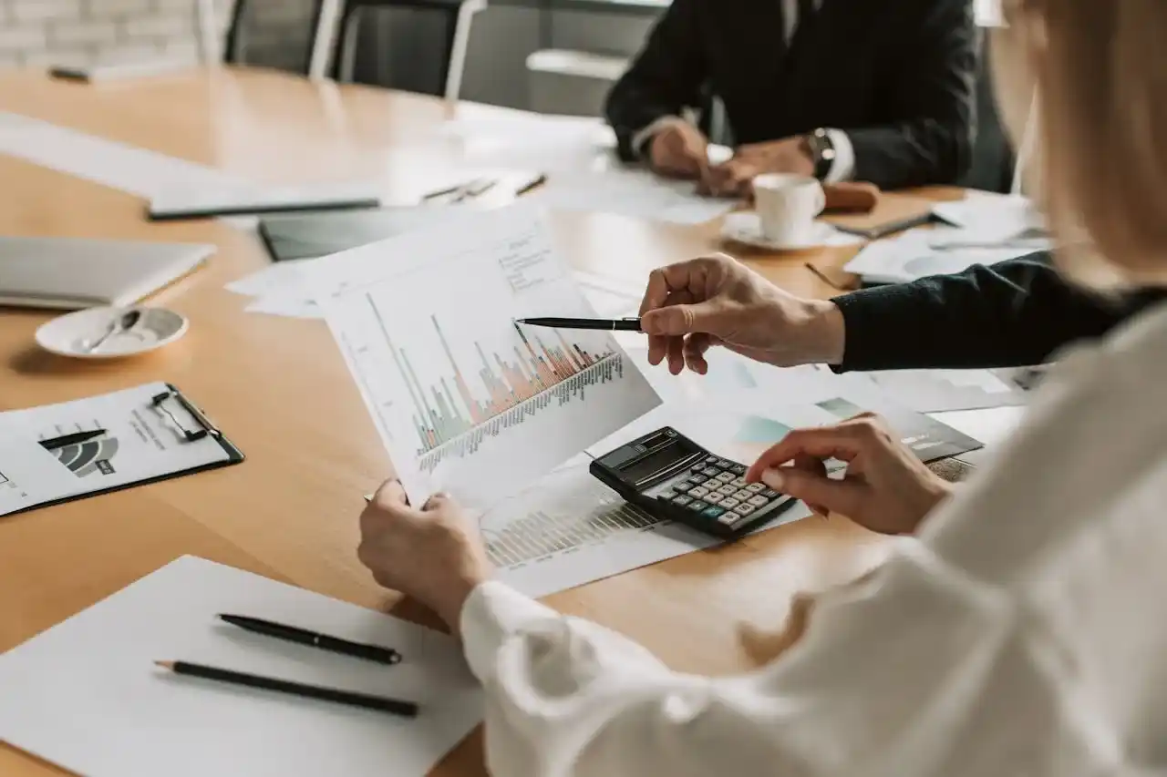 Strategic Deal Execution - A close-up shot of hands pointing at financial charts/documents on a conference table, with a laptop showing graphs visible in the frame. Include a pen, coffee cup, and glasses to add authenticity. The focus should be on the documents and hands, not faces.
